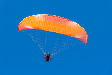 A single paraglider with a vibrant orange and red canopy is seen against a deep, clear blue sky during a sunny day flight.