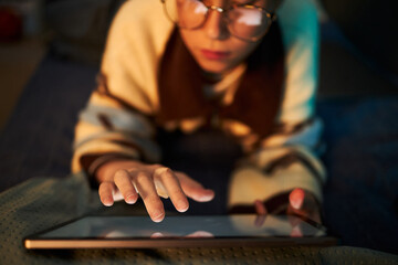 Caucasian child wearing glasses using digital tablet while lying on bed, focusing on screen with...
