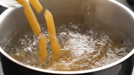 Closeup shot of dry penne pasta being carefully poured from a white bowl into a stainless steel pot filled with vigorously boiling water on a stovetop preparing a delicious Italian meal. - Powered by Adobe
