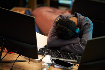 Young adult Black man wearing headphones resting head on crossed arms on desk surrounded by computer monitors, keyboard, smartphone and papers, appearing exhausted at workspace