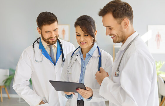 Three professional doctors standing together, looking at tablet and discussing information displayed. Smiling clinic staff in lab coats checking test results, talking about treatment in medical office