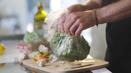 A man is holding a bag of broccoli on a cutting board. The broccoli is in a plastic bag and is being prepared for cooking