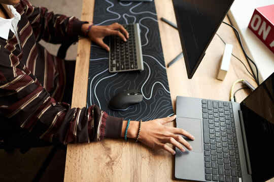 Young adult Black man working at desk using wireless keyboard and laptop, hands typing and navigating mouse, modern workspace setup with dual monitors and office accessories visible