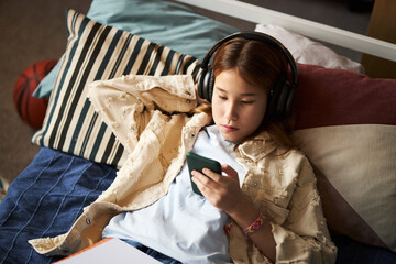 Teenage Caucasian girl lying on bed listening to music with headphones while holding smartphone,...