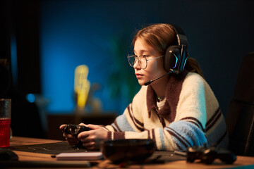 Caucasian teenage girl wearing glasses and headset playing video game with game controller, sitting at desk and focusing on screen, showing concentration and engagement in gaming