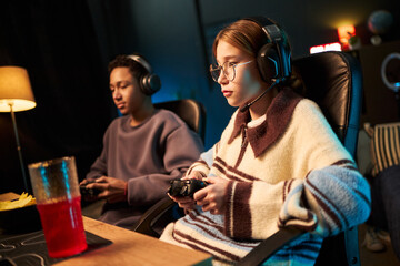 Teenage Caucasian girl and Black teenage boy, sitting in gaming chairs wearing headsets playing...