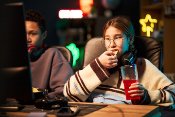 Teenage Caucasian girl wearing glasses sitting next to Black teenage boy, eating snack and drinking...