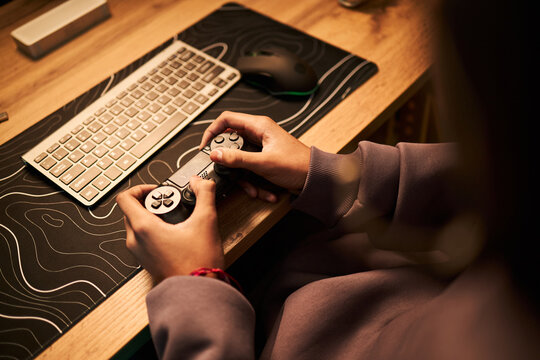 Teenager with medium skin tone holding game controller sitting at desk using computer keyboard and mouse, focusing on gaming activity, hands and partial upper body visible