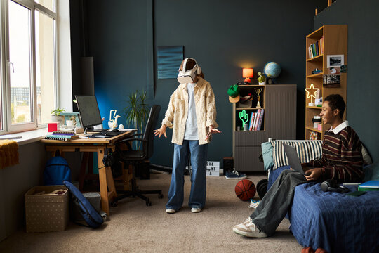 Teenage Caucasian girl wearing virtual reality headset standing in modern bedroom, gesturing with hands while teenage Black boy sitting on bed using laptop and smiling