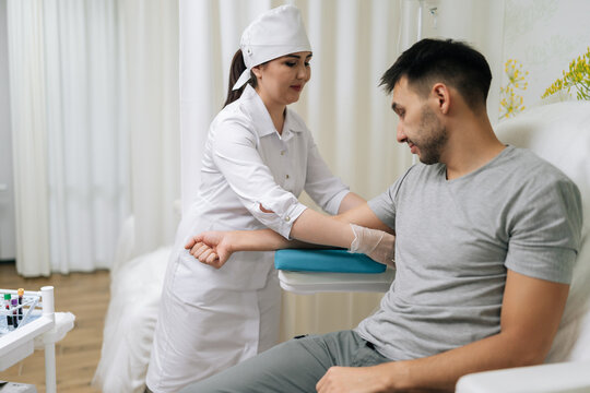 Side view of medical worker in uniform and gloves preparing equipment for drawing blood from male patient sitting in examination chair in modern clinic for health checkup.