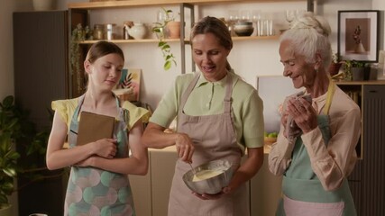 Portrait shot of grey haired grandmother in apron posing with smiling adult daughter and granddaughter while baking cookies in kitchen passing down family recipes to generations, slow motion - Powered by Adobe