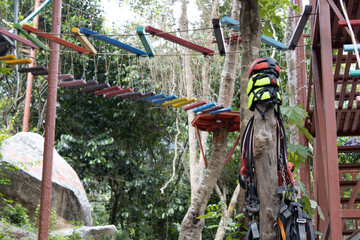 Safety gear, including a helmet and harness, hangs on a tree at an adventure park, with a colorful high ropes course visible in the lush green forest background.