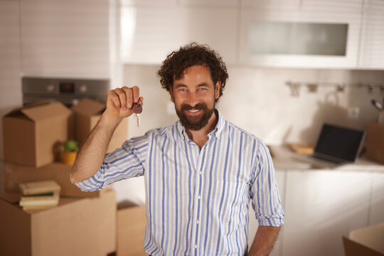 A joyful man shows house keys while standing among stacked boxes in a modern kitchen, celebrating the transition to his new home during daytime.