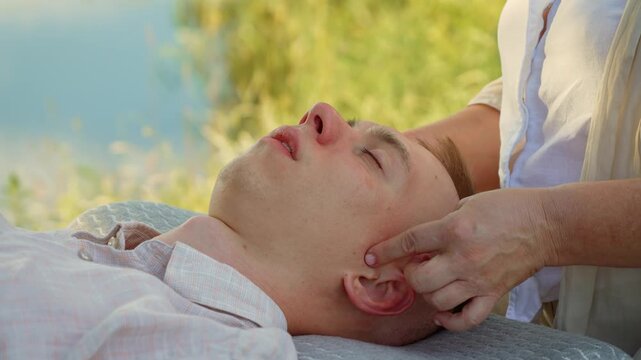 Woman massaging ear and temple of man near river for relaxation. Professional massaging head with calming energy on fresh air. Practitioner working on ear for peace and relaxation by water. Therapist