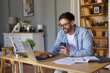 Obraz premium A cheerful man sits at a desk in a home office, typing on a laptop with a cup in hand. Surrounding him are books and a stylish lamp, creating a productive atmosphere.