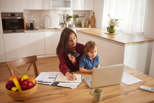 A mother helps her child with schoolwork at the kitchen table while managing tasks on her phone. They sit surrounded by family activities in a bright room. - Powered by Adobe