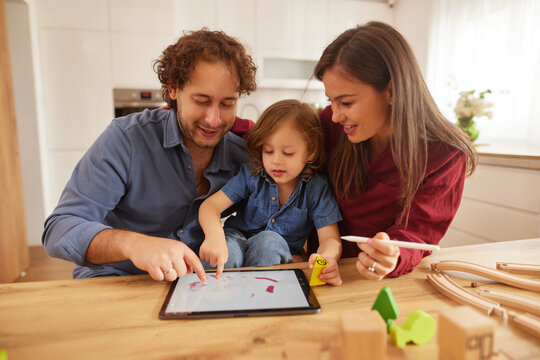 A joyful family spends quality time at home, engaging in creative play on a tablet while a young child explores colors and shapes, surrounded by toys. - Powered by Adobe
