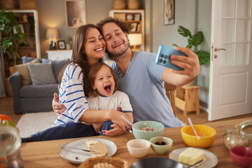 A joyful family gathers around a table filled with food. They take a playful selfie, smiling happily as they bond together at home during meal time.