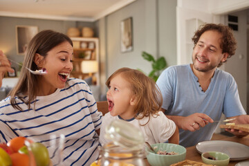 Family shares a fun mealtime filled with laughter. Parents and child express happiness while enjoying food around a dining table in a warm, inviting kitchen.
