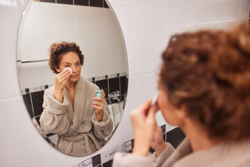 A woman with curly hair is using a cotton pad to apply a skincare product in front of a large round...