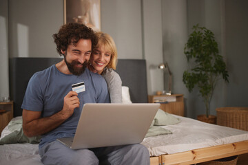 A couple happily engages in online shopping together on their bed, smiling as they hold a credit card and look at the laptop screen in a cozy bedroom setting.