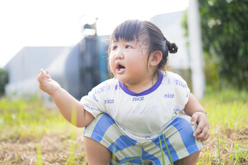A cute little Asian girl is sitting on the grass and making a funny face,Cheerful Asian girl with cute bows sitting cross-legged on green grass, enjoying a sunny day outdoors.