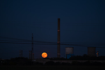 Dramatic Full Moon Rising Between Tall Industrial Chimneys and Cooling Towers at Twilight,...