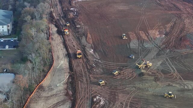 Aerial view of a vast construction site with bulldozers and excavators clearing earth, trucks hauling debris, and marked foundations outlining future massive warehouse structures.