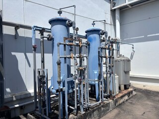 Two tall, blue industrial water filtration tanks are connected by a complex system of piping, valves, and gauges against a plain white wall. This scene illustrates the infrastructure of a commercial 