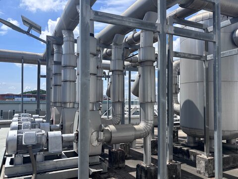 A complex view of silver industrial piping, large valves, and electric motors forming part of a chiller plant or HVAC system on a commercial rooftop. The structure shows essential mechanical infrastru