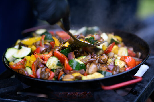 Close-up of a chef standing in a garden wearing black Nitrile gloves cooking assorted fresh vegetables in a frying pan on a gas hob