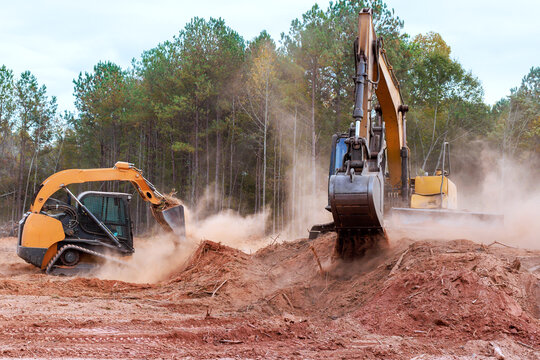 Heavy excavator machinery skid steer is working to clear dirt debris in wooded location, creating dust clouds in process.