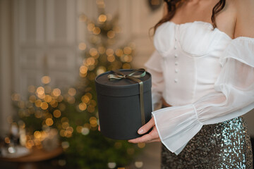 Close-up side view of a glamorous woman in a party outfit standing in front of an illuminated Christmas tree holding a gift box