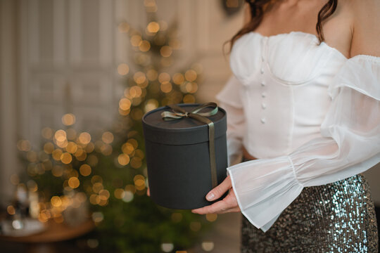 Close-up side view of a glamorous woman in a party outfit standing in front of an illuminated Christmas tree holding a gift box