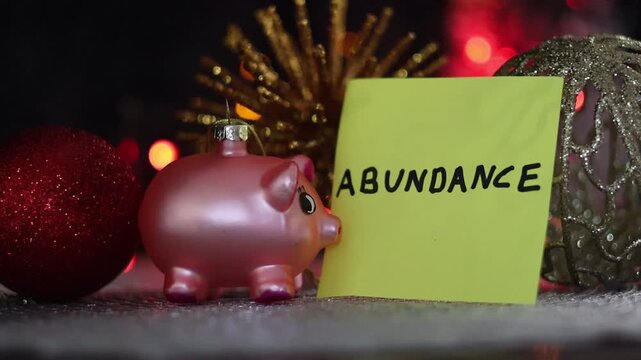 Una toma de cerca de varios adornos navide&ntilde;os y un cerdo de color rosado rodeados de luces de colores con una nota con la palabra abundancia para la familia en el nuevo a&ntilde;o. Fin de a&ntilde;o, feliz navidad