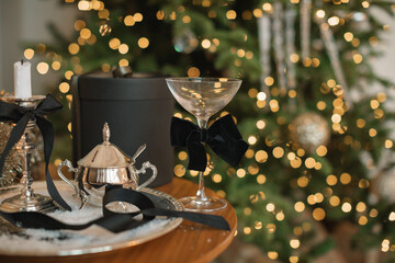 Close-up of an elegant table setting with a gift box, silverware and a champagne coupe in front of an illuminated Christmas tree