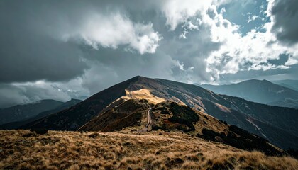 A dramatic mountain landscape features a winding path along a ridge, bathed in sunlight breaking through dark, stormy clouds.