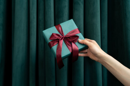 Close-up of a woman's outstretched hand in front of a green curtain holding a gift box tied with a red ribbon