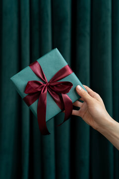 Close-up of a woman's outstretched hand in front of a green curtain holding a gift box tied with a red ribbon
