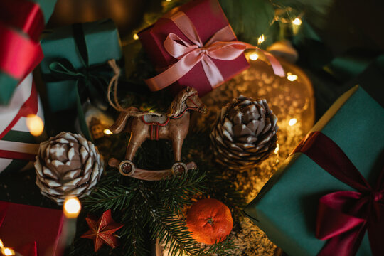 Full frame close-up of a stack of wrapped Christmas gifts, a rocking horse ornament, pinecones, fir branches and illuminated Christmas fairy lights