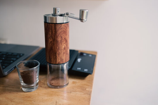 Manual coffee grinder and ground coffee in a glass on a wooden table with a laptop and scale in the background.