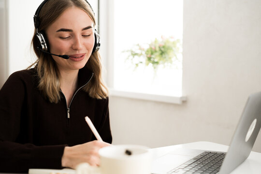 woman operator with headset writing notes in notebook working remotely in the kitchen.