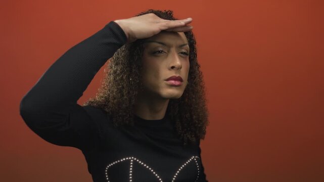 Transgender hispanic woman shading eyes with hand in studio against orange wall while wearing black studded top; curiosity.