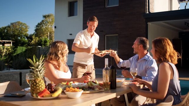 Group of adult friends having diner by swimming pool in sunny backyard.
