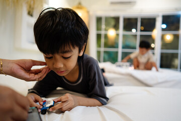 A mother gently checks on her young son while he plays with a small toy on the bed in a warm home setting. A sibling plays in the background.
