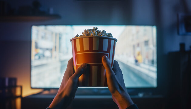 Close up on a person holding a large bucket of popcorn while watching a movie on a television at home