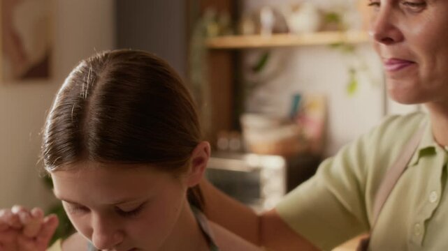 Close up of happy adult woman kissing gently teenage daughter on head while baking pastries together in kitchen, slow motion