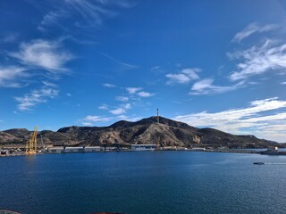 spain marcia jan 4 2024 Harbor Panorama: Hills, Cranes, and Tall Monument Rising Over Calm Blue Sea Under Clear Sky