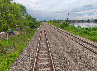 Two sets of parallel railroad tracks converging in the distance, surrounded by green trees, grass, and dry earth, with residential buildings visible in the background under a moody cloudy sky.