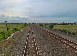 Fototapeta premium Two sets of parallel railroad tracks converging in the distance, surrounded by green trees, grass, and dry earth, with residential buildings visible in the background under a moody cloudy sky.